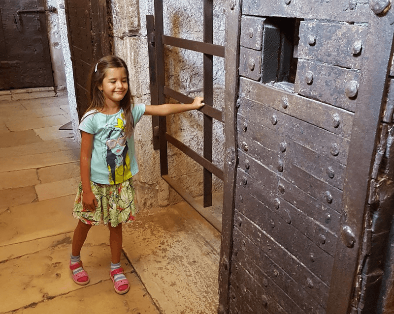 A little girl stands in the interior of an ancient building with her hands on the bars of a prison cell. 