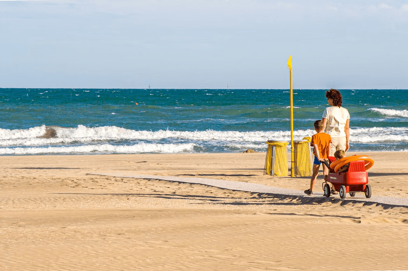 A woman and child with another child in a wagon walk on a deserted beach with the ocean in the background. 