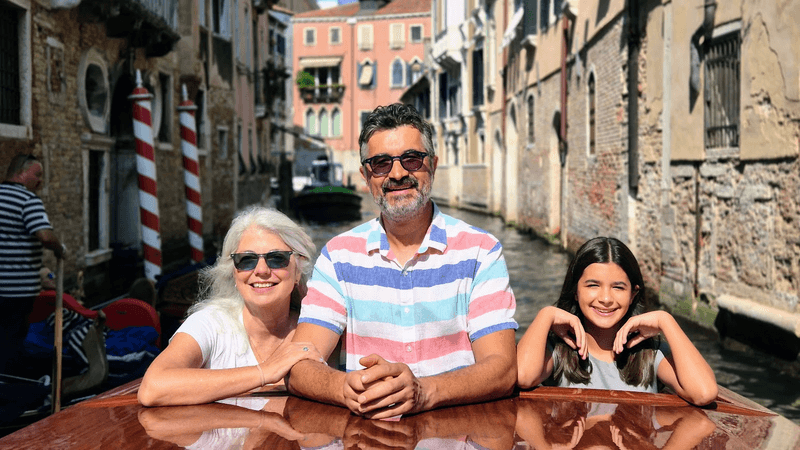 A family of two adults and a teenage girl stand in the back of a water taxi on a canal in Venice. 