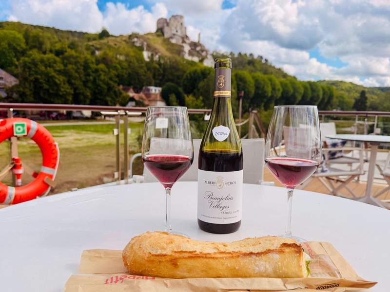 A baguette, a bottle of red wine and two glasses on the sun deck overlooking a castle in the distance.