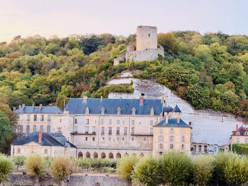 A chateau with the ruins of a castle above it as seen from the river.