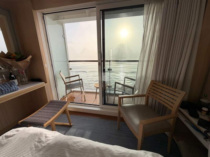 A bed, a chair, a small bench, in a river cruise cabin with a sliding glass door and veranda.