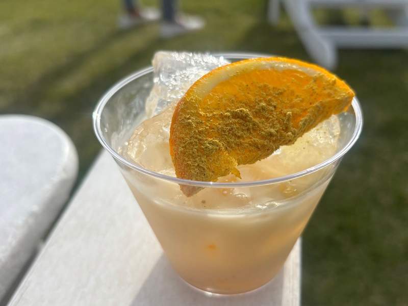 A clear plastic cup filled with an iced drink, garnished with a powdered orange slice, sits on a white surface outdoors.