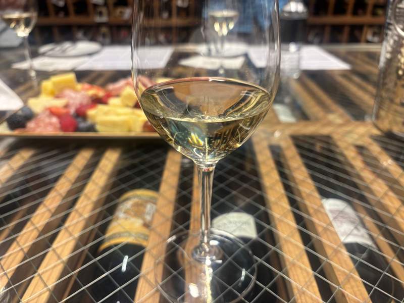 A close-up view of a wine glass filled with white wine, sitting on a table with visible wine bottles underneath and a charcuterie board in the blurred background.