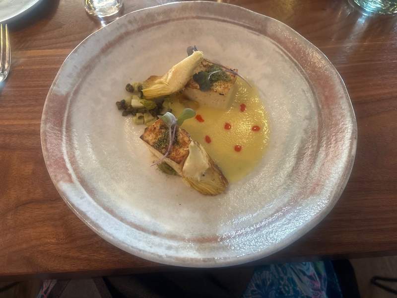 A gourmet plated dish of seared protein served with artichoke hearts, capers, a yellow sauce, and red garnish dots on a wide, textured white bowl.