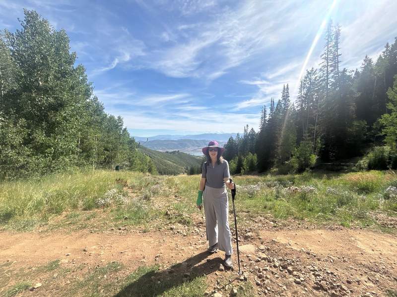 A person wearing a hat, sunglasses, and light-colored hiking attire stands on a dirt trail with trekking poles, surrounded by grassy fields, trees, and mountain views in the background.