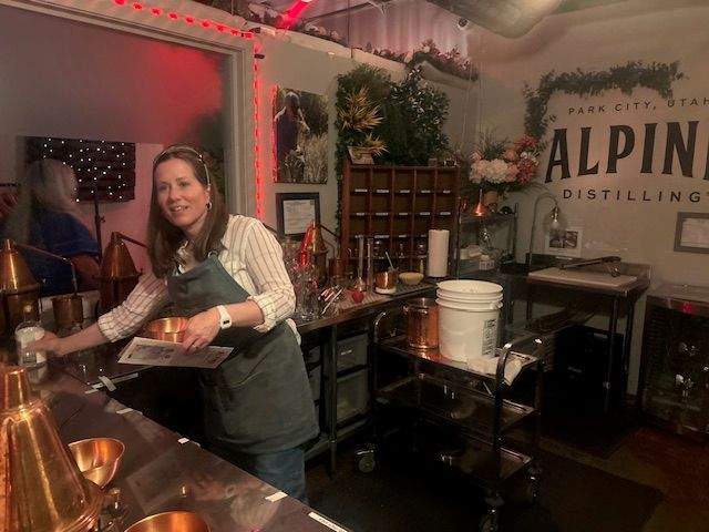 A woman wearing a striped shirt and a green apron works behind a counter at Alpine Distilling, surrounded by distillery equipment and a sign on the wall