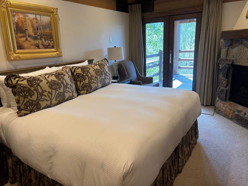 A hotel bedroom featuring a large bed with white linens and patterned throw pillows, a painting on the wall, an armchair by a balcony door, and a stone fireplace.