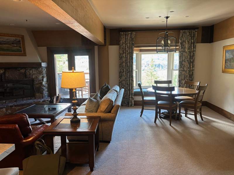 A spacious living and dining area with a sofa, coffee table, stone fireplace, and a dining table with chairs next to a large bay window.