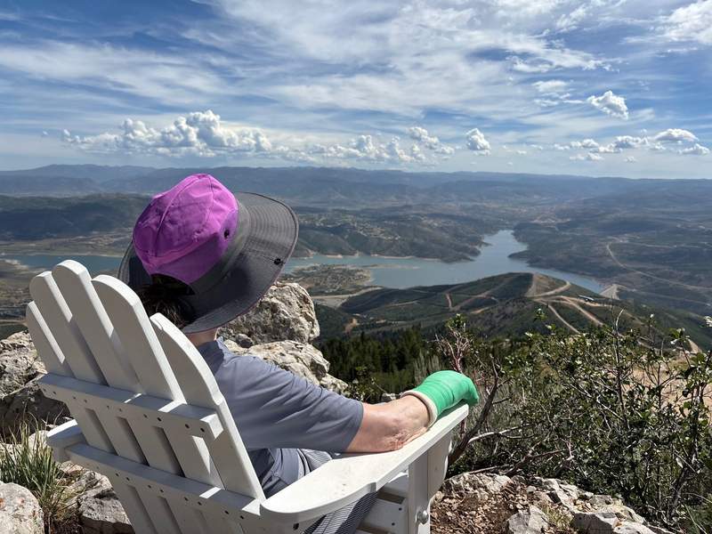 A person wearing a purple and grey sun hat sits in a white Adirondack chair on a rocky overlook, looking out at a scenic view of a large body of water and distant mountains.