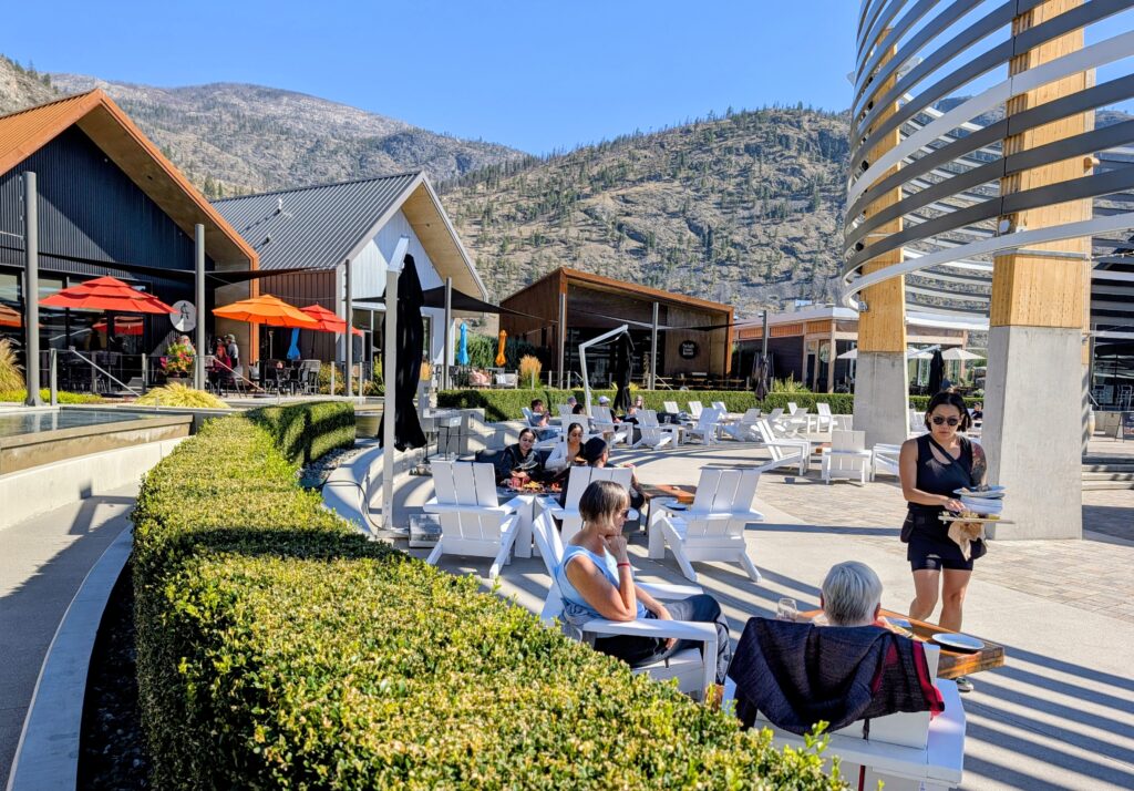 Woamn waiting on peopel sitting at tables in the sunshine rnged by buildings that house boutique wineries at the District Wine Village in Canada's Osoyoos Wine Region