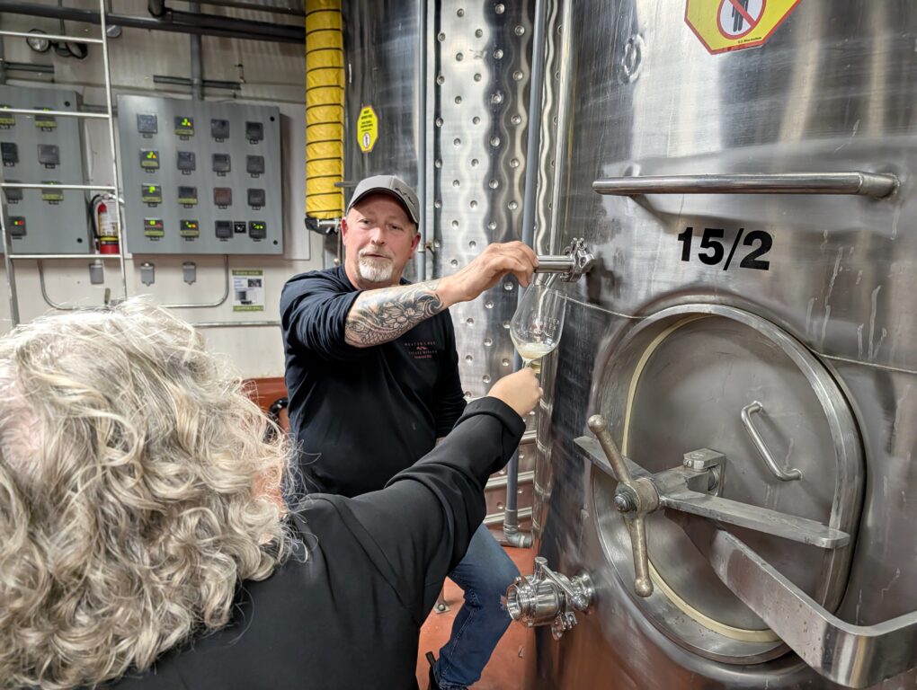 Man pours a glass of Chardonnay for a woman hold a glass up to the spigot of a stainless steel fermentation tank