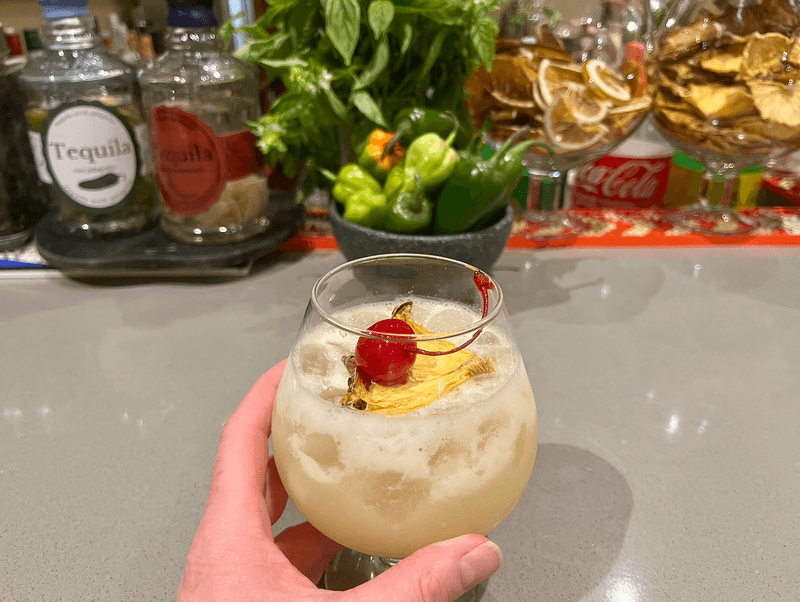 a hand holding a tropical cocktail garnished with a cherry with liquor bottles in the background