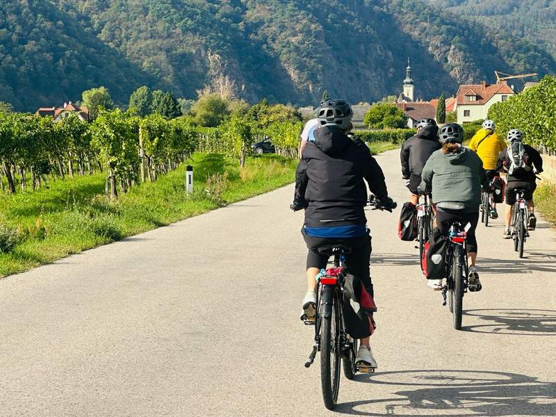 Cyclists ride past vineyards and a small village surrounded by forested hills in Austria's Wachau Valley.