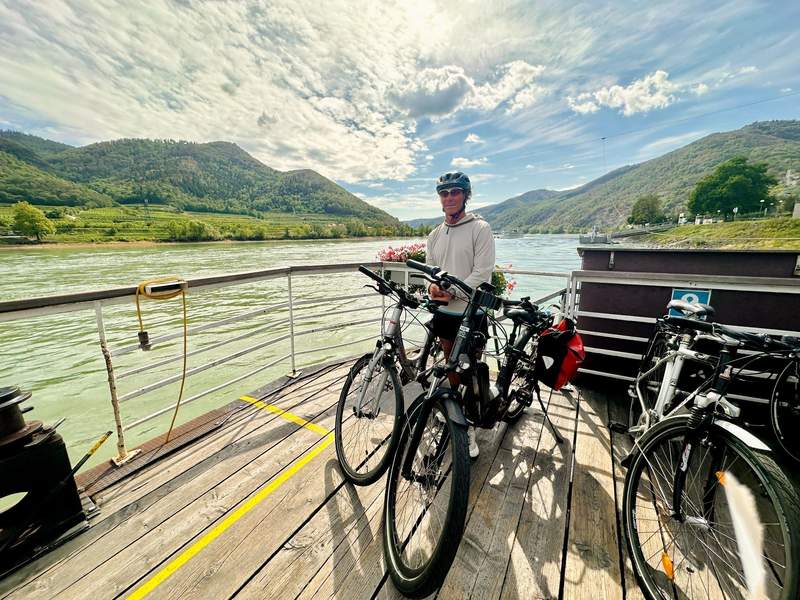 A cyclist wearing a helmet stands with touring bikes aboard a ferry crossing the Danube, with forested hills and vineyards in the background.