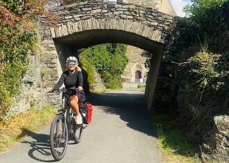 A cyclist wearing a helmet rides an e-bike beneath a stone archway on a quiet paved path.