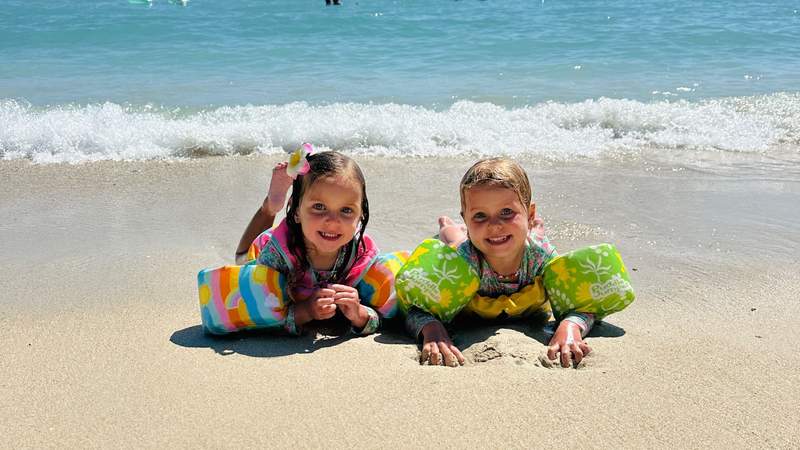 Two young girls wearing colorful floaties smile on a sandy beach with turquoise Hawaiian water behind them.