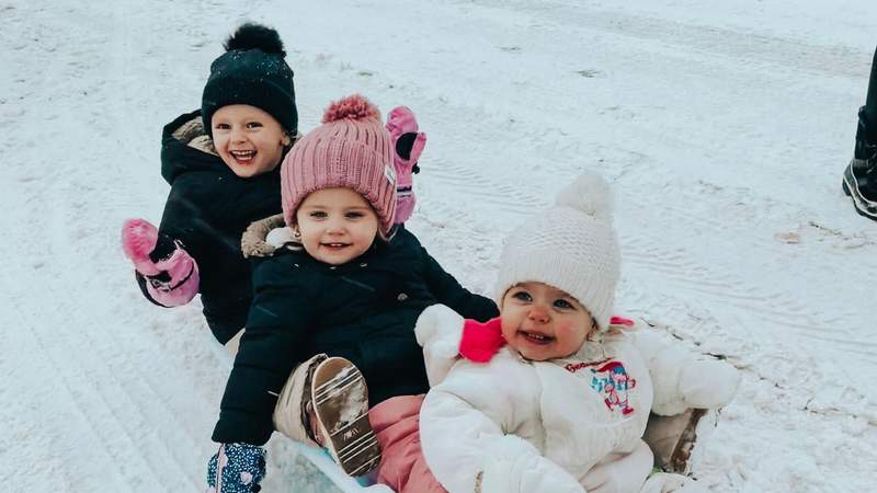 Three young girls bundled in winter coats and knit hats smile together while playing in the snow.