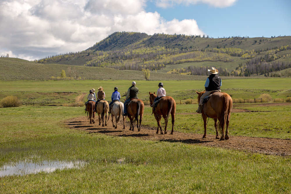 A group of horseback riders follows a dirt trail through green meadows at C Lazy U Ranch in Colorado.