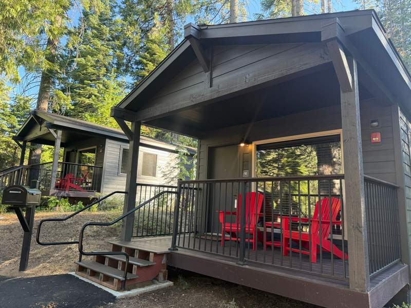 Dark gray cabin at Waybound Yosemite surrounded by tall evergreen trees, featuring a covered front porch with two bright red Adirondack chairs.