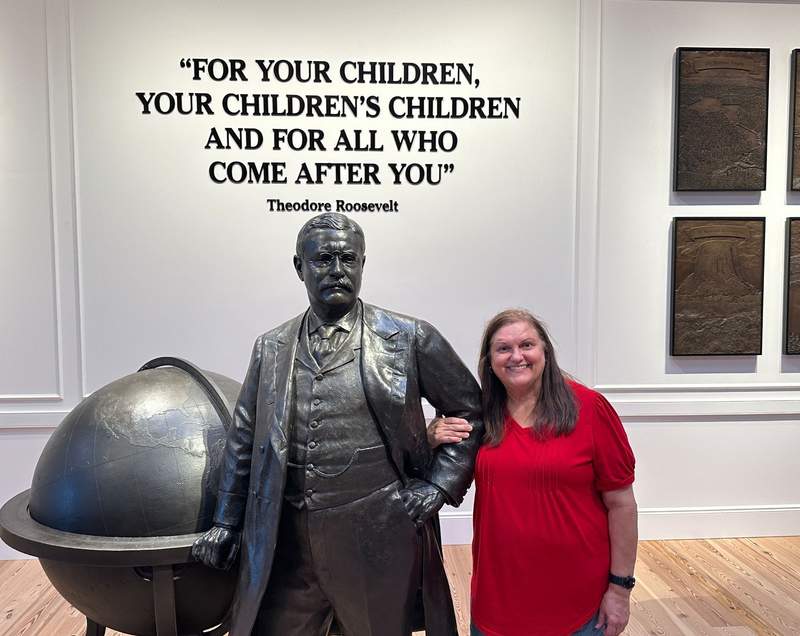 Lady in a red top standing beside a Theodore Roosevelt Statue in the Presidential Museum.