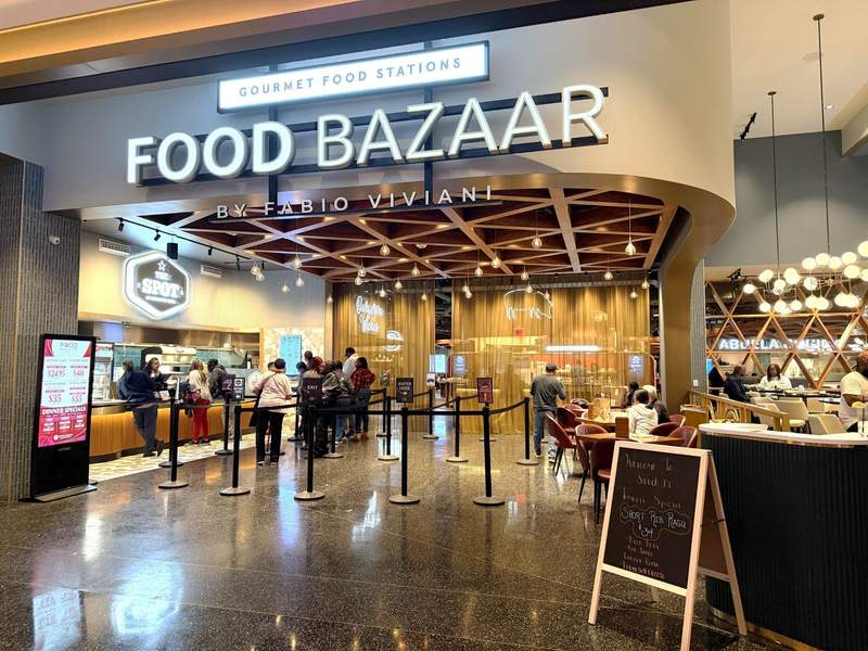 Entrance sign and dining area for Food Bazaar by Fabio Viviani at Wind Creek Chicago Southland including people waiting in line at The Spot outside the bazaar.