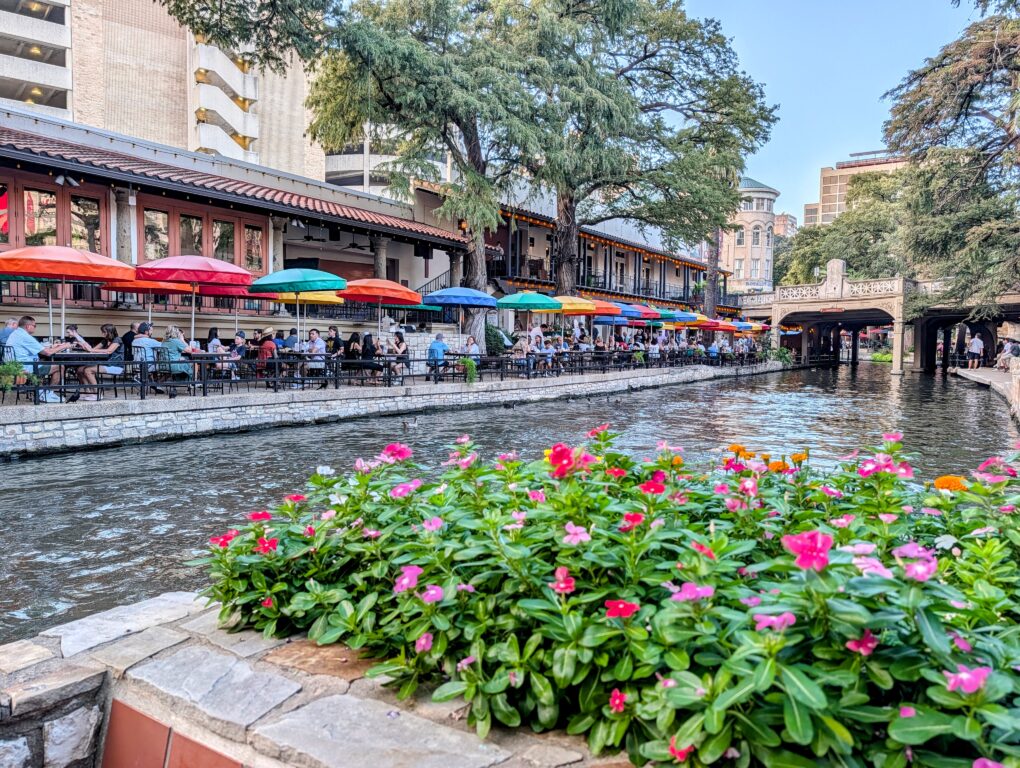 San Antonio River Walk, with flowers on one side of the river and colorful umbrellas over riverside tables on the other