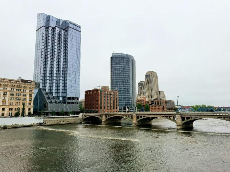Downtown Grand Rapids skyline along the Grand River, featuring the Amway Grand Plaza tower, modern high-rise buildings, and a bridge crossing the water. 