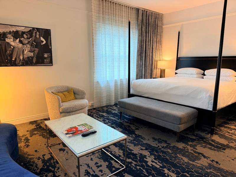 Stylish guest room at Amway Grand Plaza featuring a four-poster bed, upholstered bench, marble-top coffee table, comfortable seating, and framed artwork.