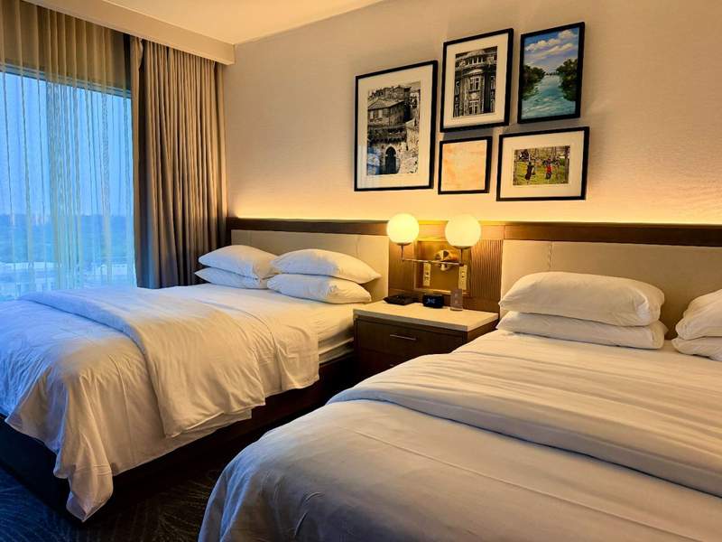Two-queen guest room at JW Marriott Grand Rapids featuring white bedding, modern bedside lighting, framed artwork and a large window with sheer curtains.