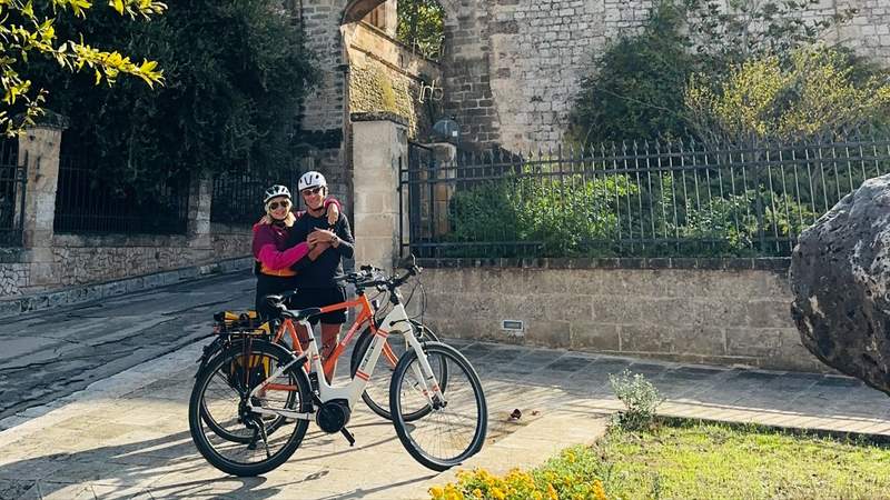 A couple wearing cycling helmets pose together beside their rental bicycles on a historic stone street in Puglia Italy