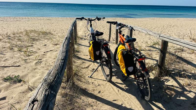 Two touring bicycles with yellow pannier bags parked beside a wooden beach walkway overlooking a sandy beach and turquoise sea in Puglia Italy