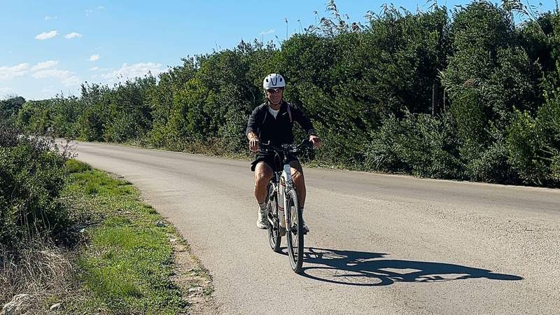 A man wearing a bicycle helmet rides a touring bike along a quiet paved road surrounded by green vegetation in Puglia, Italy.