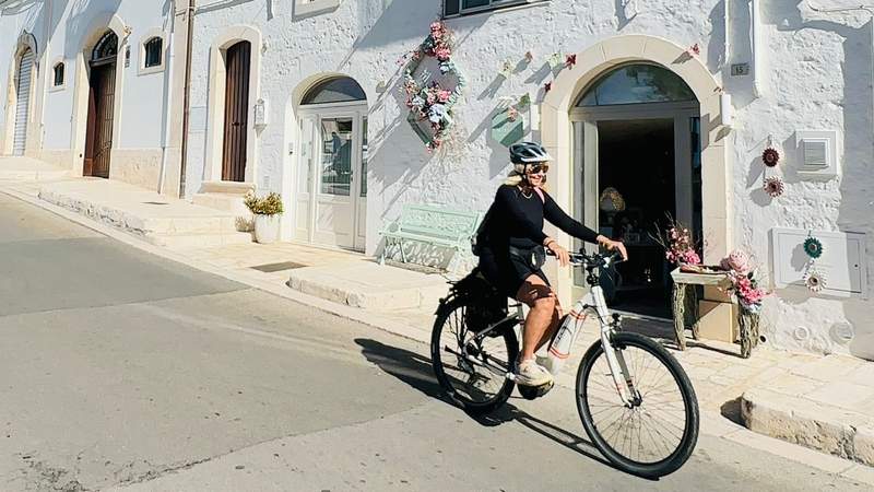 Woman wearing a helmet rides an e-bike past whitewashed buildings and flower-decorated storefronts in Alberobello, Puglia, Italy.
