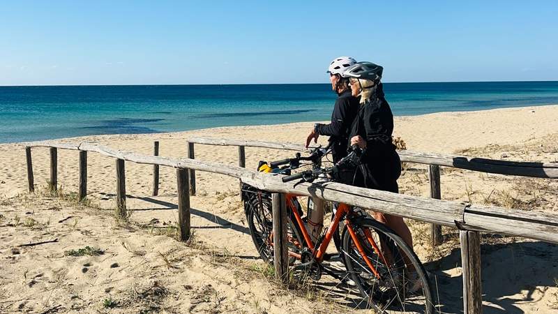 A couple wearing cycling helmets stand beside their bicycles overlooking a quiet sandy beach and turquoise Mediterranean Sea in Puglia, Italy.