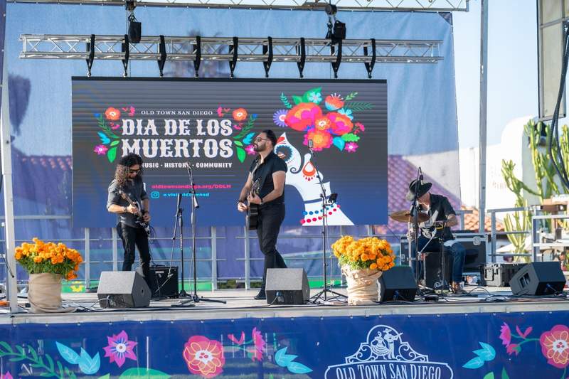 Three musicians performing on the Old Town San Diego Dia de los Muertos stage flanked by buckets of orange marigolds