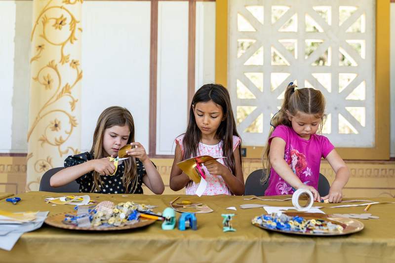 Three young girls cutting gold paper and decorating crafts with sequins at a table in front of painted columns