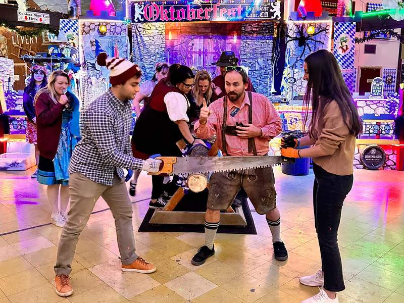 Two contestants pull a crosscut saw through a log as a host in lederhosen coaches them at Oktoberfest in Big Bear Lake, California